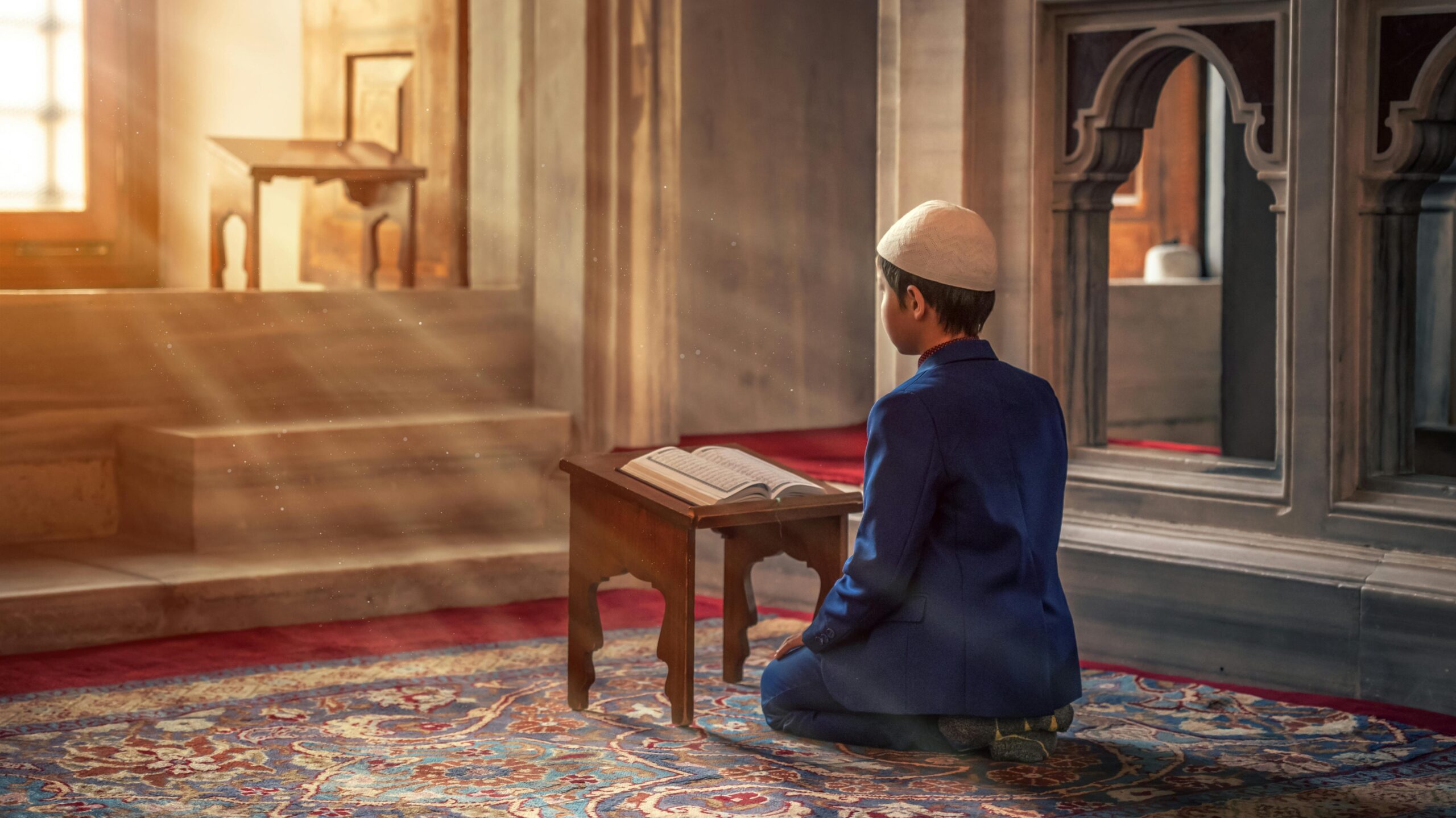 A young boy kneels in prayer inside a mosque with sunlight streaming through the window.