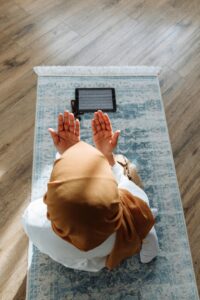 A woman in a hijab prays on a rug with a tablet nearby, in a peaceful indoor setting.
